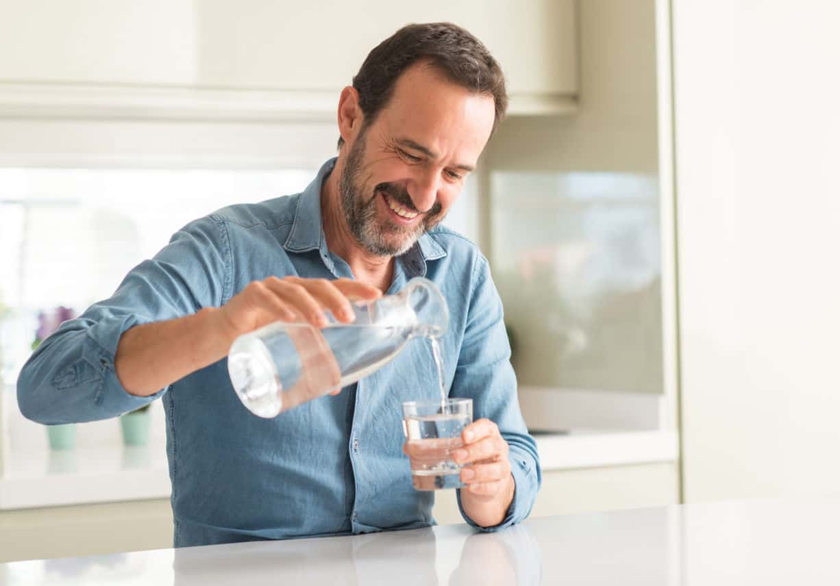Middle age man drinking a glass of water with a happy face standing and smiling with a confident smile showing teeth