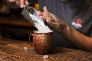 person-pouring-ice-cubes-into-moscow-mule-cup-on-wooden-ba