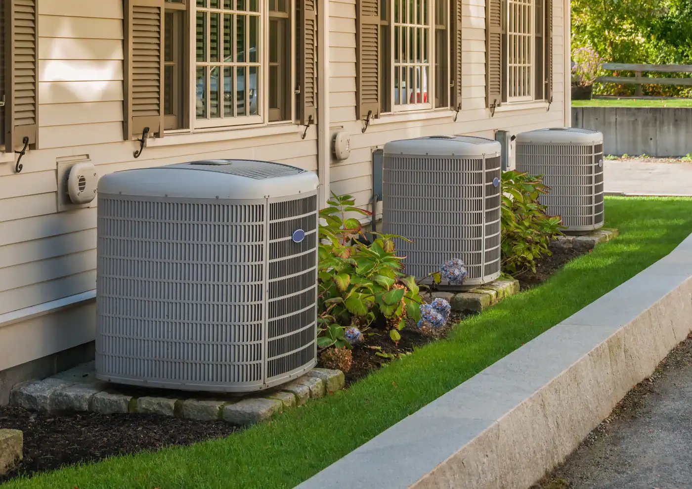 Three outdoor air conditioning units are lined up against the side of a beige house with brown shutters. They are set on concrete slabs surrounded by neatly trimmed grass and small flowering plants. A walkway curves alongside.