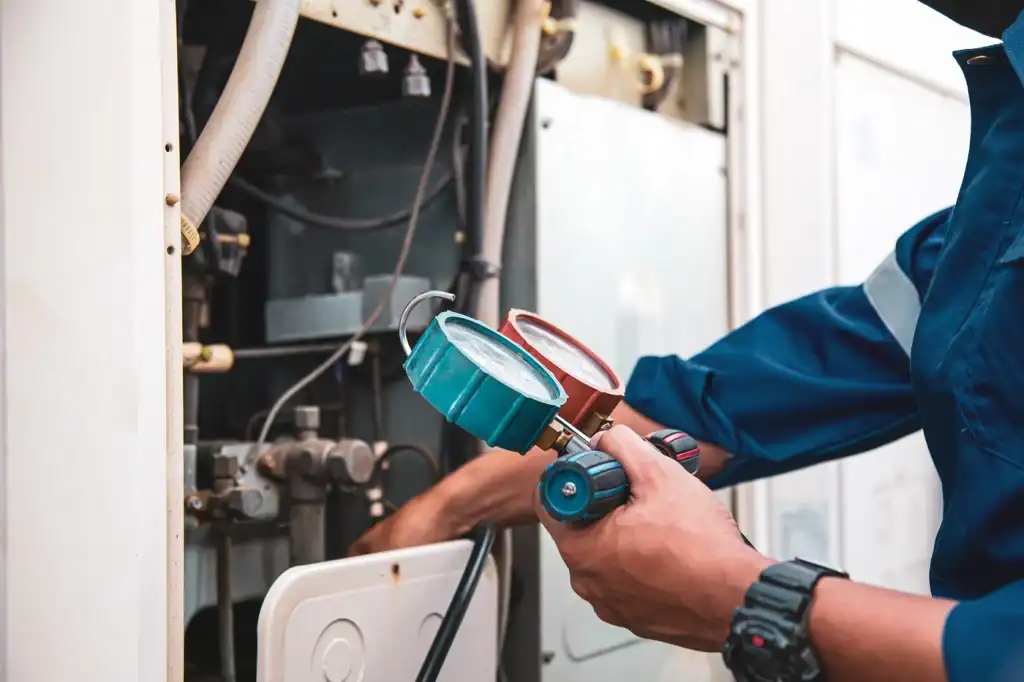 A technician in a blue uniform checks pressure gauges while servicing industrial equipment, focusing on tubes and controls. His hands hold blue and red marked gauges, with other tools visible in the background.