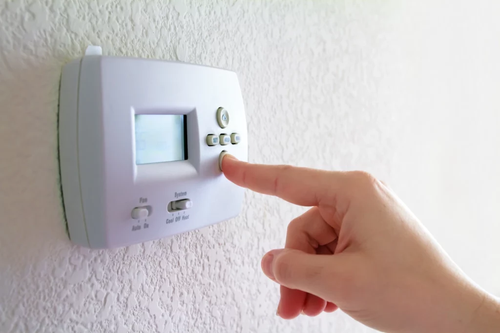 A hand adjusting the temperature on a white digital thermostat mounted on a textured wall.