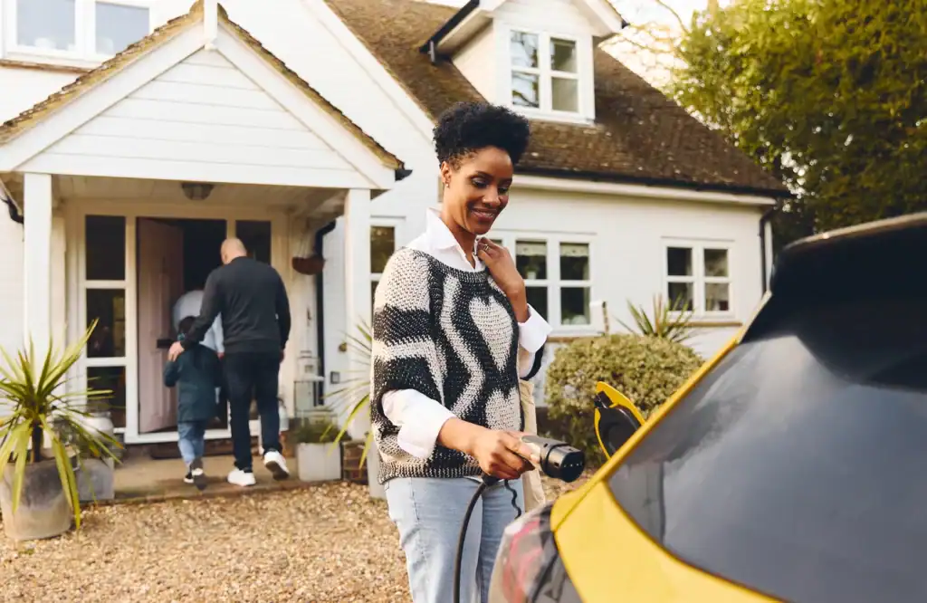 A woman stands in front of a house, smiling as she plugs a charging cable into a yellow electric car. In the background, a man and child walk towards the front door. The house is white with a wooden porch.