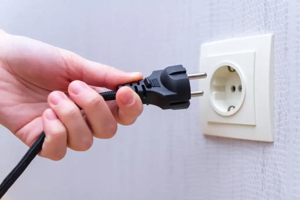 A hand grips a black power plug near a white electrical outlet on a light-colored wall, poised to connect it. The scene highlights the action of plugging in the device, an essential step to make your home energy-efficient and save on utility bills.
