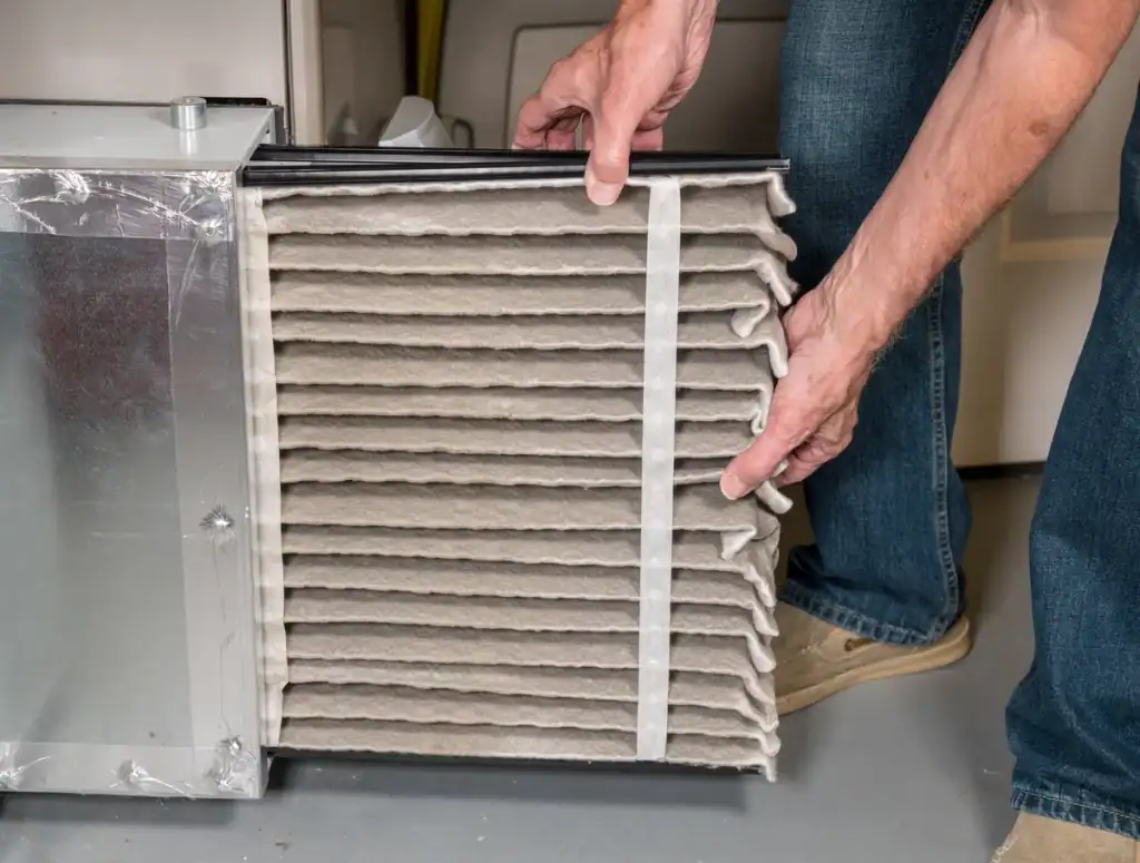 A person, possibly noting one of the "7 Signs It's Time for a New Furnace," is removing a dirty air filter from an HVAC system. The rectangular filter, framed in metal, is visibly dusty. Wearing jeans and beige shoes, they stand on a gray floor as they tackle this essential maintenance task.