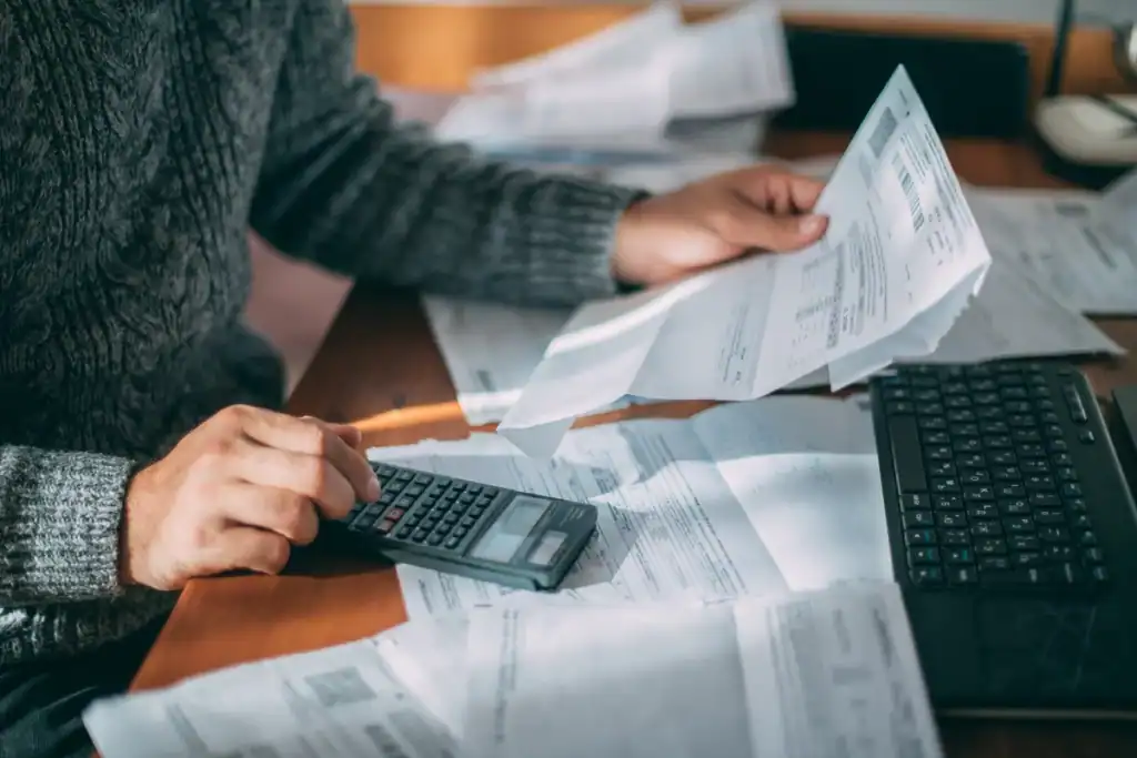 A person in a gray sweater intently uses a calculator, holding a sheet of paper amidst documents on a wooden table, with an open laptop nearby. The scene hints at making your home energy-efficient to save on utility bills, reflecting careful financial planning or bill management.