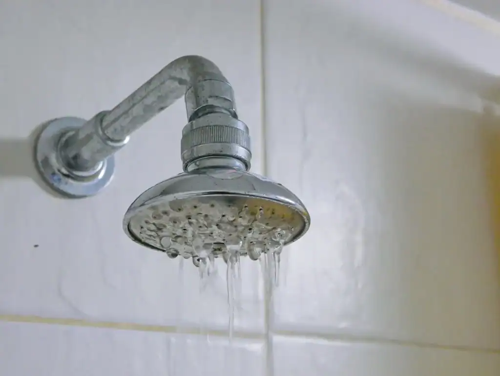 A metal showerhead mounted on a tiled bathroom wall with water flowing from it in multiple streams. The white wall tiles are pristine, suggesting a recent installation or DIY project, though the showerhead appears slightly worn from use.