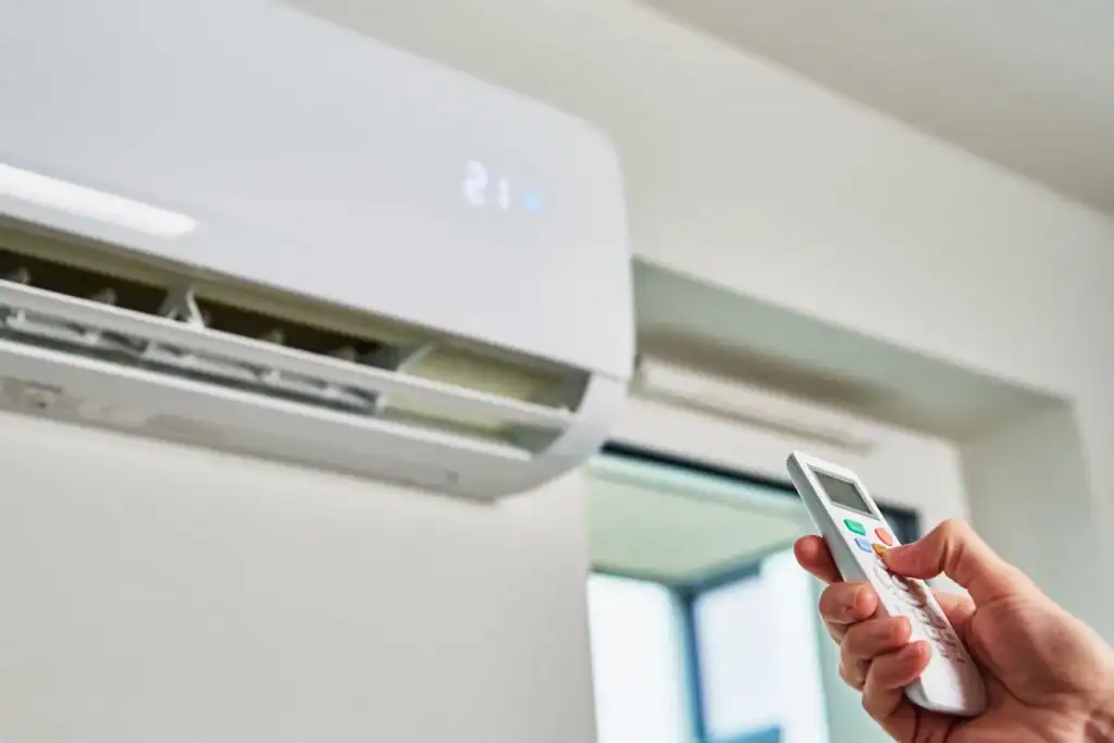 A hand holding a remote control is pointed towards a white wall-mounted air conditioning unit displaying a temperature of 21°C. The unit, likely installed by one of the top HVAC companies in Minneapolis, is positioned above a window and appears to be operating efficiently.