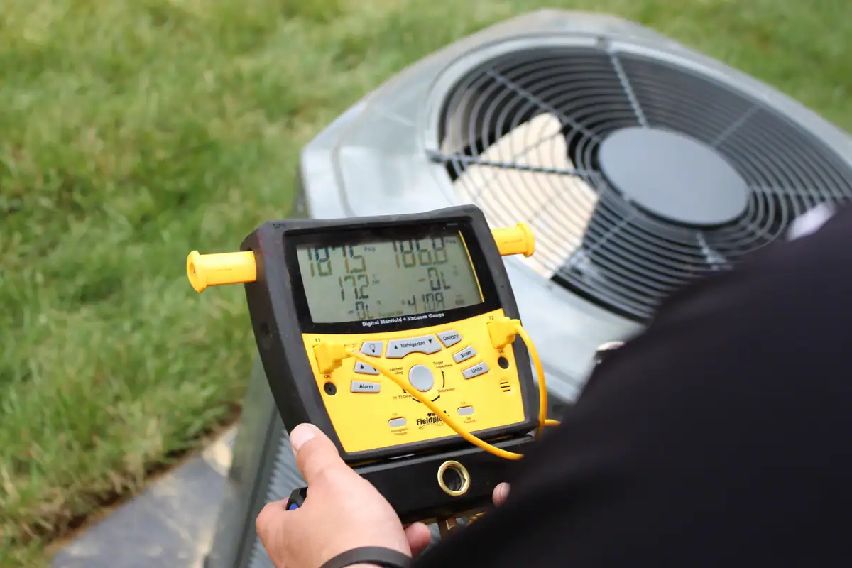 A person holds a yellow electronic device displaying numbers in front of an outdoor air conditioning unit. The digital manifold gauge measures various HVAC system parameters, a tool commonly used by HVAC companies in Woodbury. Green grass is visible in the background.