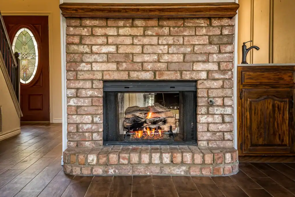 A cozy indoor fireplace with a red brick surround and a glass door. Logs are burning inside, creating a warm glow. The fireplace is set against a backdrop of wooden floors and adjacent cabinetry. An ornate, oval-shaped glass door is visible in the background.