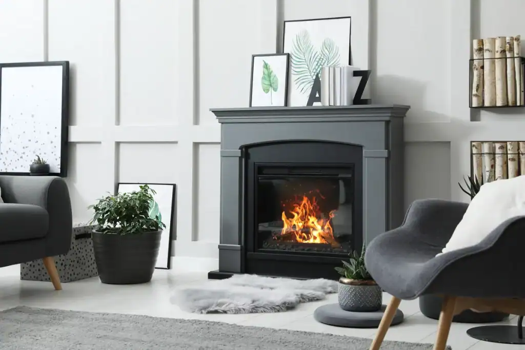 A modern living room with a lit fireplace, surrounded by decor such as framed leaf prints, books, and plants. It features grey armchairs, a grey and white rug, and a black fireplace with a fur rug in front. The walls are white with rectangular panel designs.