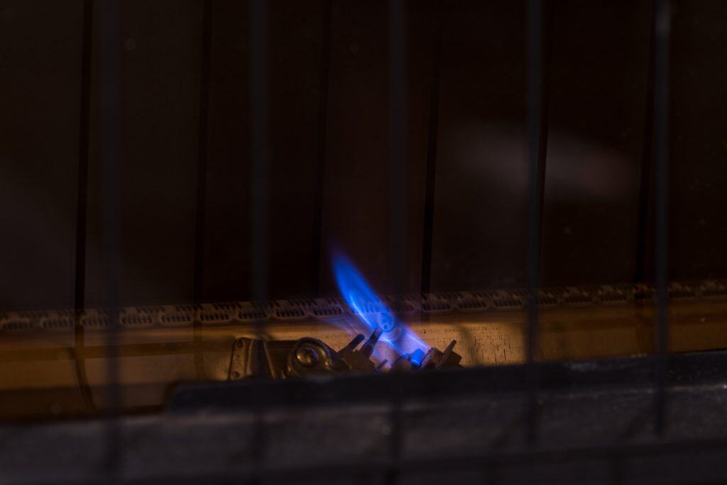 A close-up of a small blue flame burning inside a metal grate, reminiscent of a furnace pilot light that won't stay lit. The flame emerges from a gas burner, casting a subtle glow on the surrounding dark surface while the grate bars are visible in the foreground.