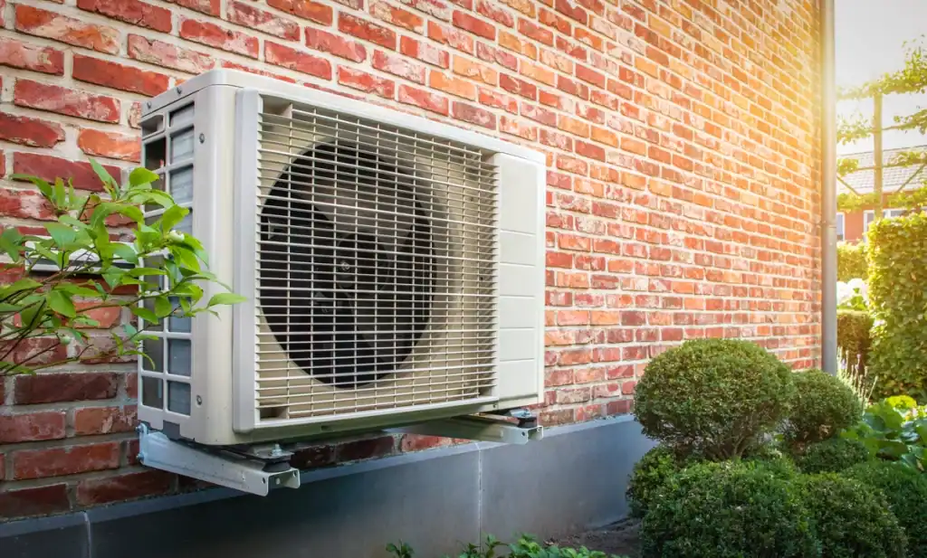 An outdoor air conditioning unit, possibly resembling a heat pump, is mounted on a brick wall with lush greenery, including bushes and plants, in the foreground. Sunlight gently illuminates the scene from the right, highlighting nature's calm amidst technology.