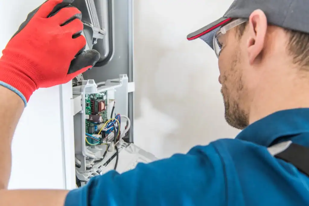 A technician wearing a cap, safety glasses, a blue shirt, and red gloves is examining and repairing an open electrical panel with visible wiring and components. The side perspective focuses on the technician's hands and the internal parts of the unit, raising questions like "how long does a furnace last" due to detailed inspections.