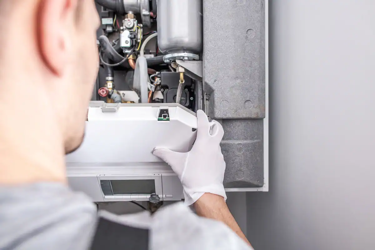 A technician wearing white gloves is inspecting and adjusting parts inside an open boiler or HVAC system against a light gray wall. The focus is on the internal components and the technician's hands, skillfully manipulating a panel, as if determining how long a furnace lasts through meticulous care.