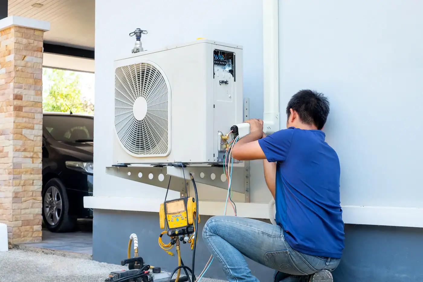 A technician in a blue shirt is expertly installing or repairing an air conditioning unit on the building's exterior wall, demonstrating how to install a heat pump. Various tools and equipment are scattered beside him, while a car peeks out from under the carport in the background.