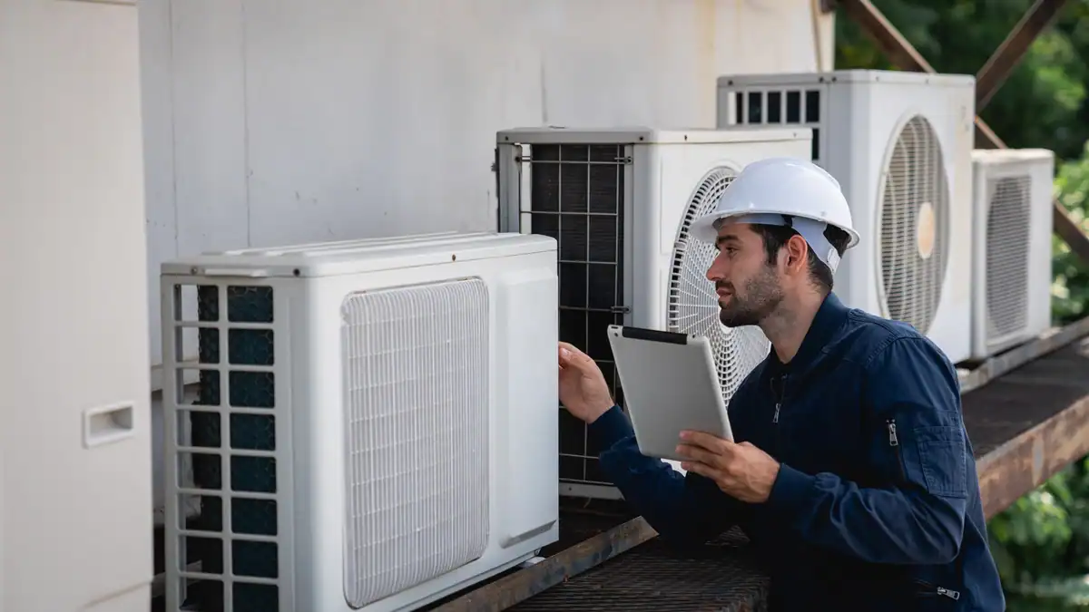 A technician in a white hard hat and blue uniform inspects an air conditioning unit on a rooftop, possibly evaluating the heat pump installation cost. He holds a tablet for reference while examining the equipment, with several other AC units mounted in the background.