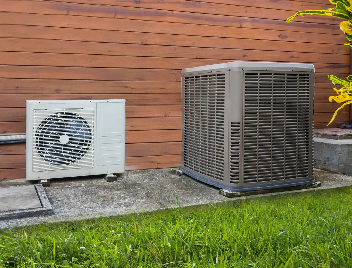 Outdoor air conditioning units next to a wooden wall, surrounded by grass. One unit is white with a visible fan, and the other is larger and gray with a grid pattern—both designed to maximize heat pump efficiency.