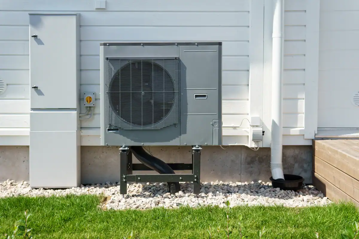 An outdoor heat pump is installed on a white house wall, situated on a metal stand above a bed of small stones. Green grass surrounds the area, and a white downspout is visible to the right, illustrating an efficient setup without adding significant installation costs.