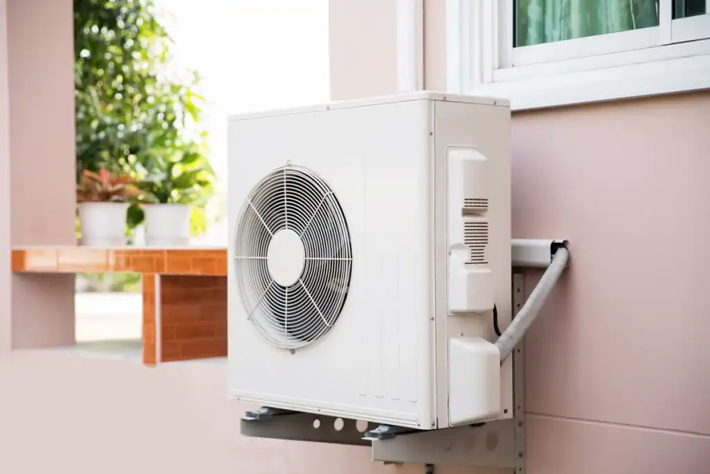 A white outdoor air conditioning unit, resembling a heat pump, is mounted on a wall beside a window. The unit is connected by cables and has a visible fan. In the background, potted plants and greenery add a touch of nature.