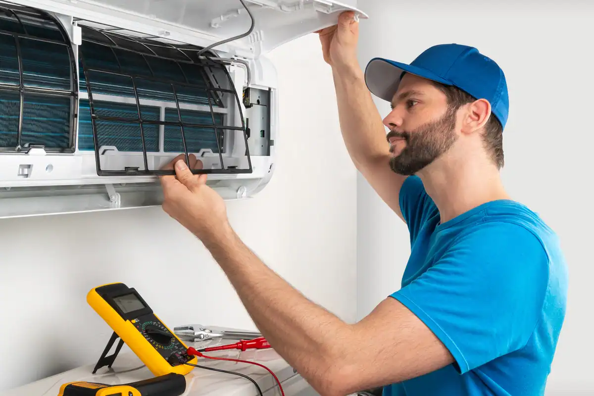 A technician in a blue shirt and cap expertly adjusts the front panel of an air conditioning unit. As a multimeter and wires rest below, the well-lit room highlights his skillful focus on maintenance—a crucial step in managing the cost to repair an air conditioner.