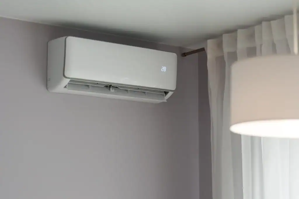 A modern white air conditioning unit is mounted on a gray wall, boasting a sleek design as it displays 28 degrees. With sheer white curtains beside it and the edge of a white lampshade in view, you might wonder about the cost to repair an air conditioner if needed.