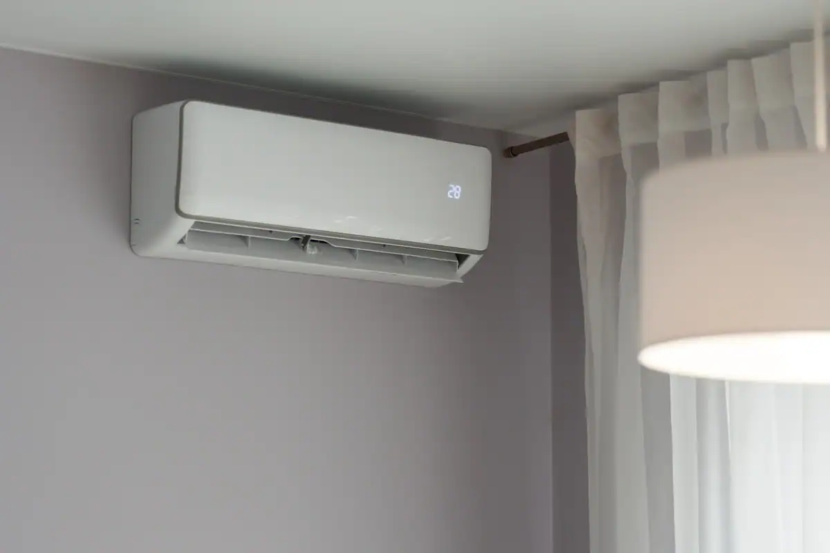 A modern white air conditioning unit is mounted on a gray wall, boasting a sleek design as it displays 28 degrees. With sheer white curtains beside it and the edge of a white lampshade in view, you might wonder about the cost to repair an air conditioner if needed.