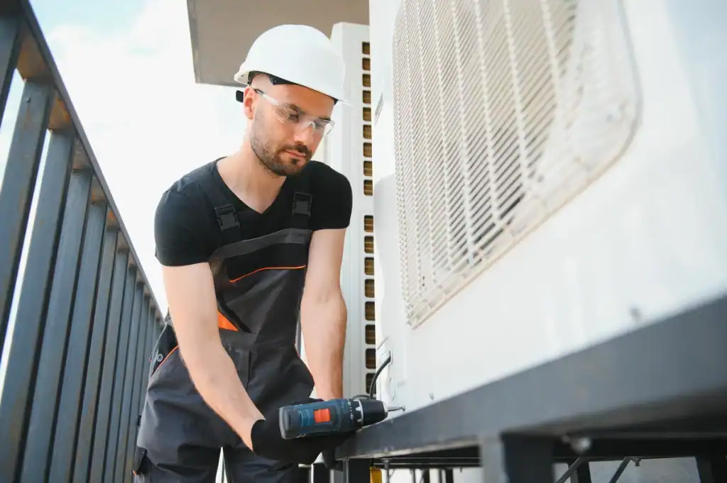 A technician wearing a white hard hat and safety goggles expertly conducts heat pump maintenance on an outdoor air conditioning unit. Dressed in dark work overalls, the technician operates a power drill while standing on the balcony.