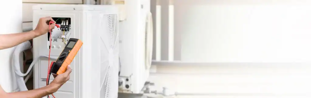 Person using a digital multimeter to check the electrical connections of an outdoor air conditioning unit. The person is holding the device against the open compartment, examining parts of a heat pump, while blurred background elements suggest an exterior setting.