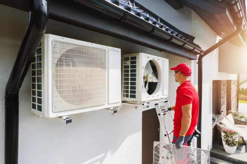 A technician in a red uniform and cap works on an air conditioning unit that doubles as a heat pump not heating, mounted on a house exterior. He stands on a ladder, holding a panel. It's a sunny day, and the units are beneath a black rain gutter.