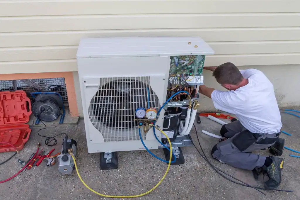 A technician kneels on the ground, repairing an outdoor air conditioning unit that isn't functioning properly. Tools and equipment are scattered around as they work on the exposed wires and components of the heat pump, all set against a beige wall.