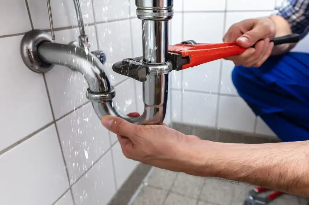 A person using a red pipe wrench to fix a leaking metal pipe under the sink, with water spraying from the joint. Clad in blue trousers, they wonder, "How much does a plumber cost?" The scene unfolds against white tiled walls.
