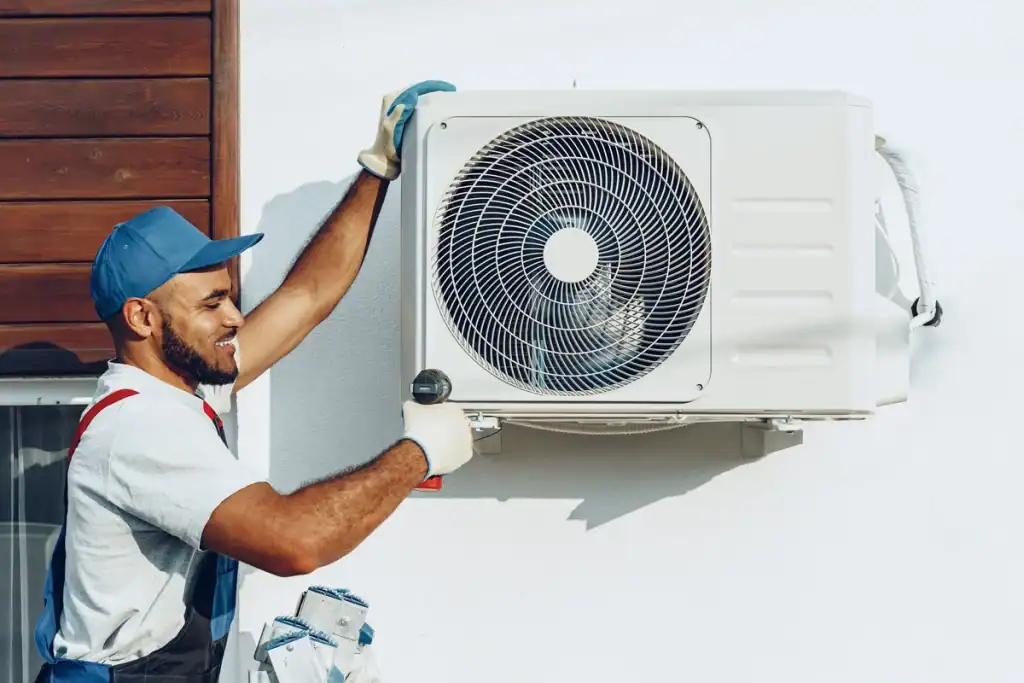 A technician in a blue cap and overalls is working on an outdoor air conditioning unit attached to a wall. With a smile, he uses a wrench, perhaps contemplating how long it takes to replace an AC unit. A wooden panel is partially visible on the left.