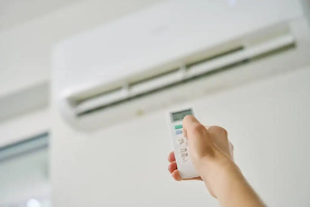 A hand holding a remote control is pointed towards a white wall-mounted air conditioning unit, perhaps contemplating how long it takes to replace an AC unit. The remote's display is faintly visible, with the room's background softly out of focus.