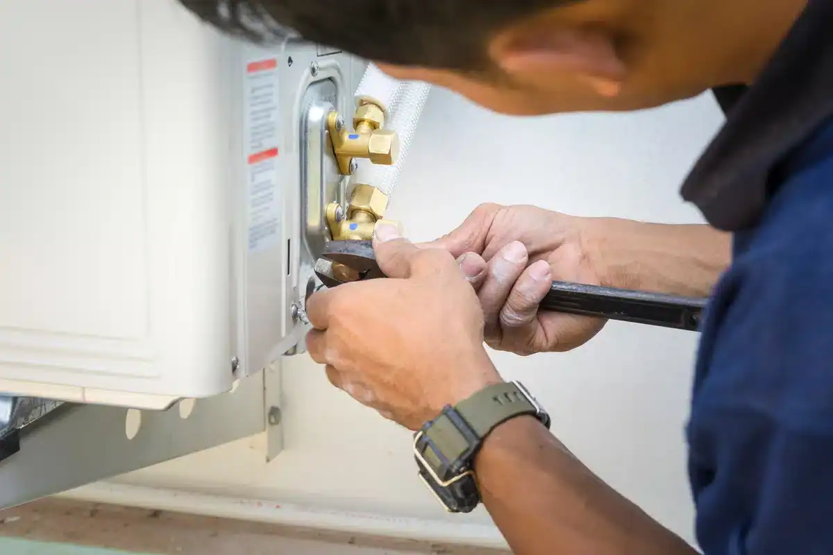 A person wearing a wristwatch and navy blue shirt uses a wrench to adjust or install a component on an outdoor air conditioning unit mounted on a wall. Their hands are securely gripping the wrench, showcasing their expertise in air conditioning repair as they work on the metallic fittings of the unit.