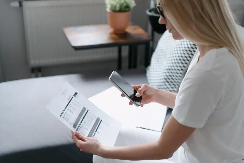A woman in a white outfit sits on a couch, using her smartphone to scan a document. A table with a small plant adds to the cozy atmosphere, creating an ideal setting for researching how to save on water bills in this home or casual office environment.