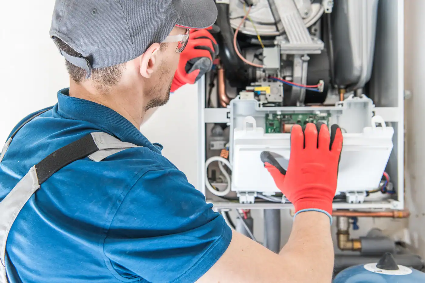 A technician wearing a cap and gloves is focused on a boiler repair, meticulously adjusting components inside the open panel with a tool. Wiring and parts of the system are visible as he follows his furnace tune-up checklist in the well-lit space.