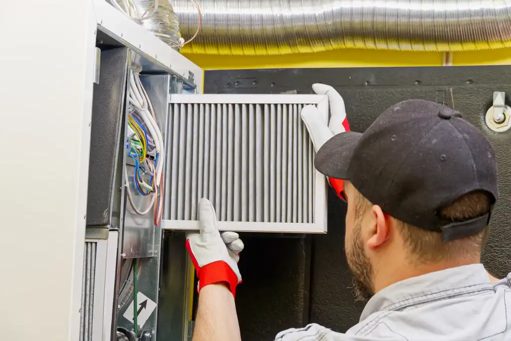 A person wearing gloves and a cap is installing or removing a pleated air filter from a ventilation system, perhaps pondering how often you should service your furnace. Wires are visible inside the unit, and metal ductwork is seen above.