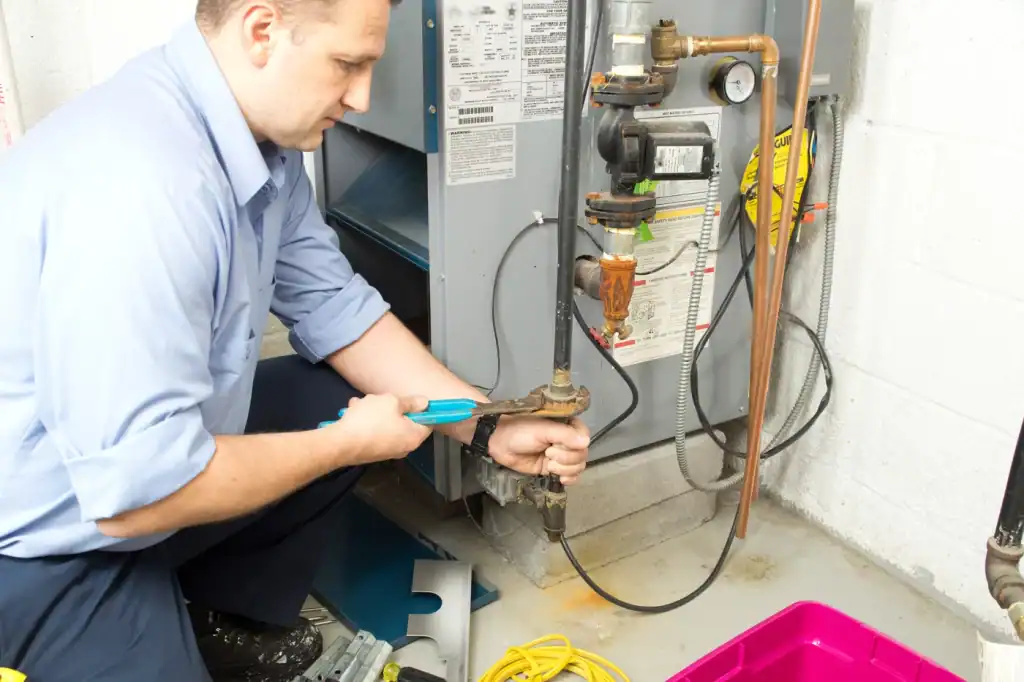 In a utility room, a person in a blue shirt uses a wrench to adjust a valve on plumbing connected to an appliance. Surrounded by tools, a yellow cord, and a pink container, they consider how often you should service your furnace for optimal performance.