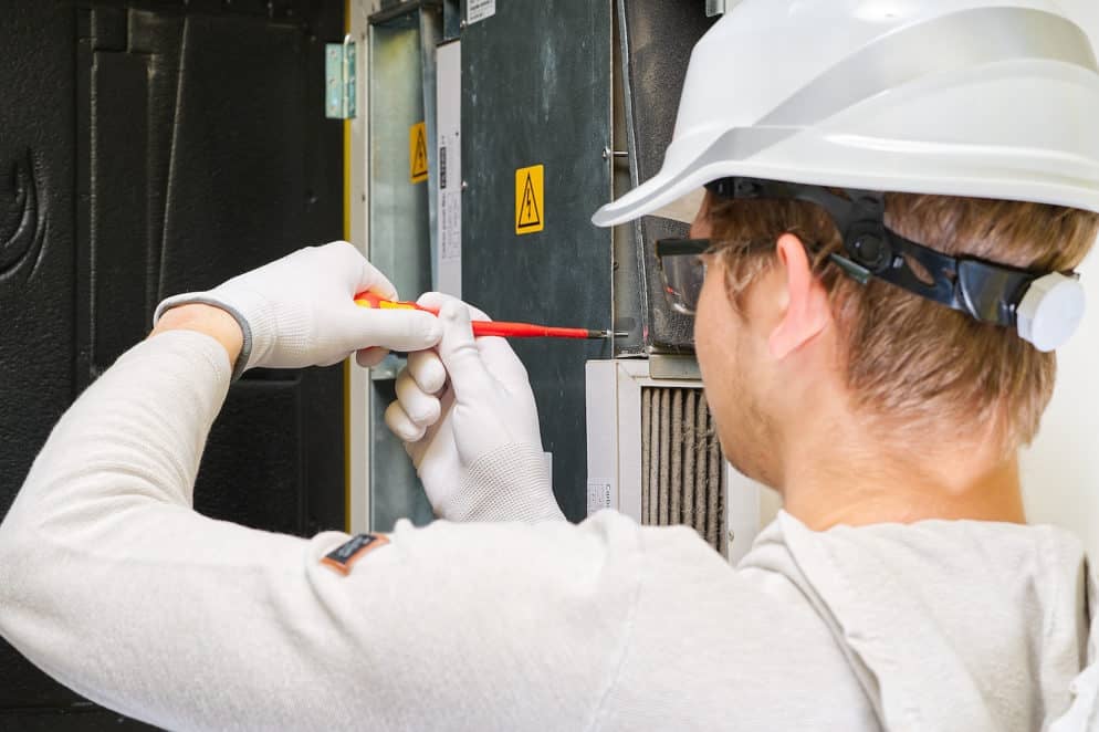 A person wearing a white safety helmet, white gloves, and a light-colored hoodie uses a red screwdriver to work on the internal components of an industrial machine. Yellow caution signs are visible on the machine's surface, suggesting HVAC service is underway.