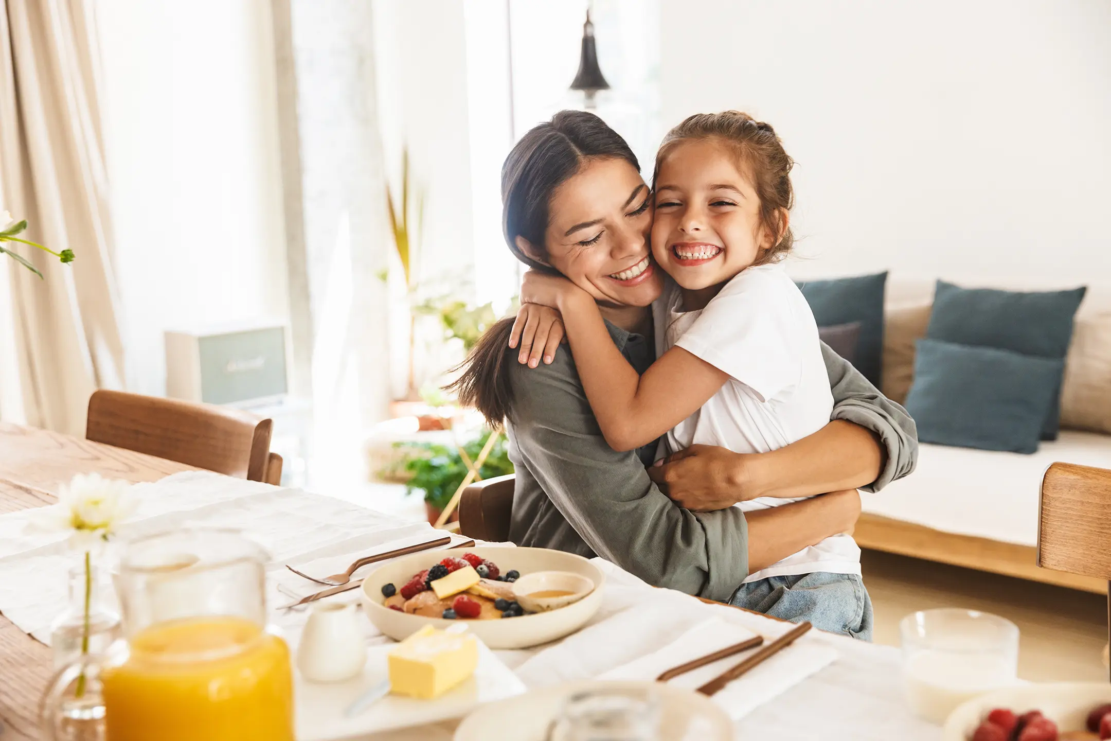 A woman and a young girl share a warm hug while seated at a wooden table set for breakfast. On the table are a bowl of fruit, a glass of juice, butter, and other breakfast items. Both are smiling joyfully in their bright, cozy dining room after discussing plans to call an electrician.