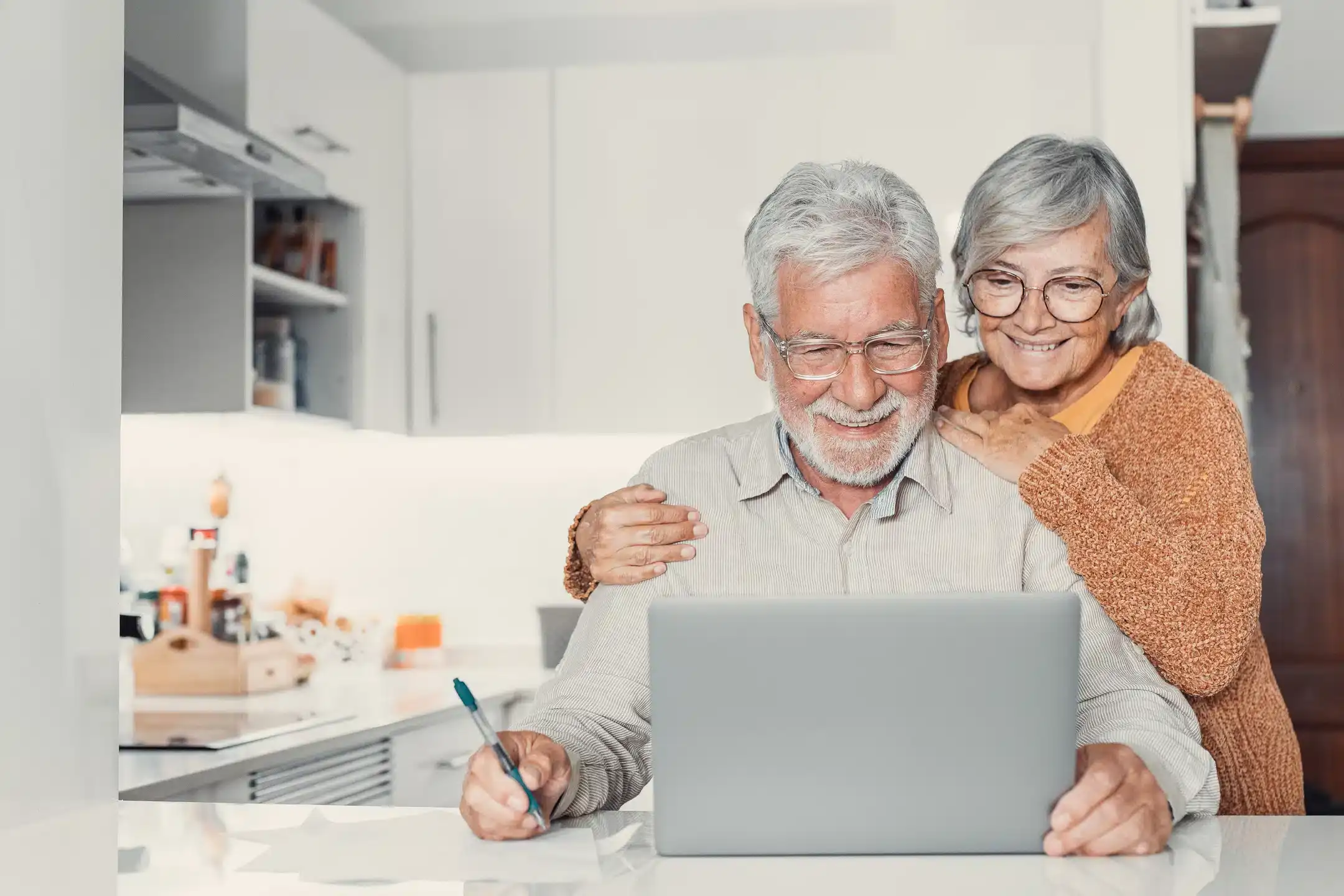 An elderly couple is in a modern kitchen, smiling as they look at a laptop screen. The man, holding a pen, sits in front of the laptop while the woman, in glasses and a cozy sweater, stands behind him with her hands on his shoulders. They seem thrilled after scheduling their furnace repair online.