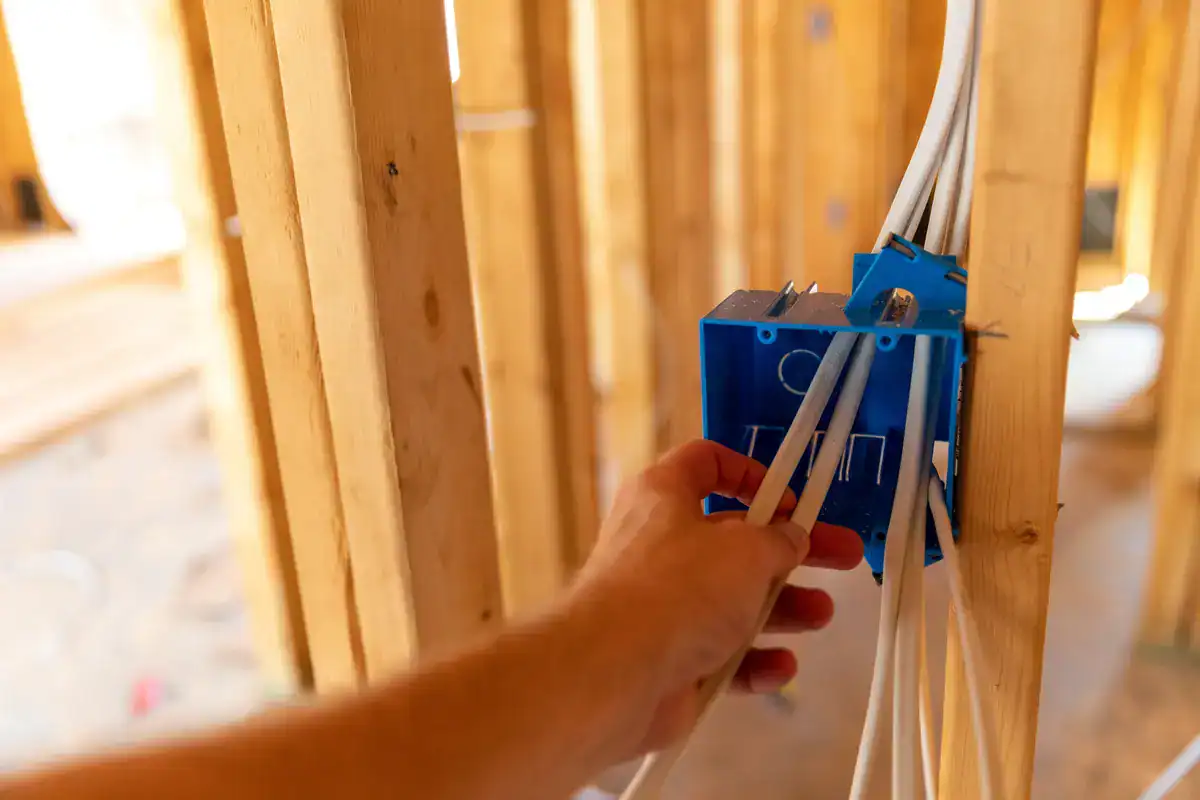 A person is installing electrical wires into a blue plastic junction box in a wooden frame structure of a house under construction. The image shows close-up details of the wiring and the wood studs, perfect for anyone searching for an electrician near me (Brooklyn Park).