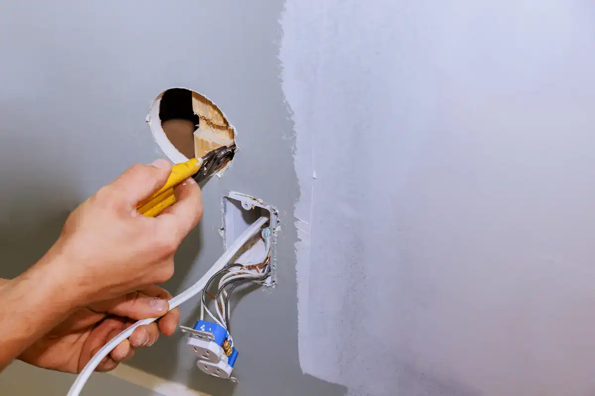 A person uses a pair of pliers to strip the insulation off electrical wires in a wall. There are exposed wires coming out of a hole in the wall, with a patch of fresh paint around the hole. The scene appears to be part of a home renovation or repair project by an electrician near me (Coon Rapids).