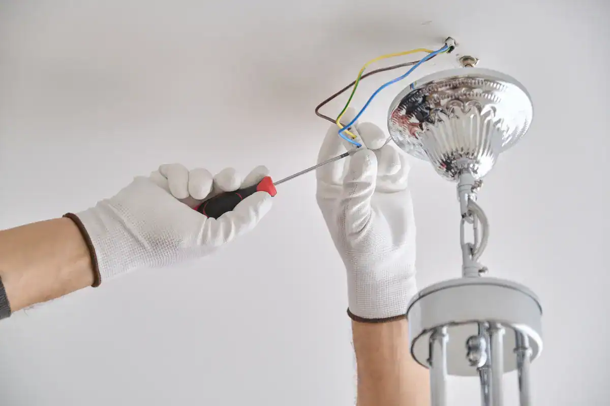 An electrician near me, wearing white gloves, uses a screwdriver to install or repair a ceiling light fixture. Several wires protrude from the ceiling, connecting to the light fixture with its metallic, reflective base.