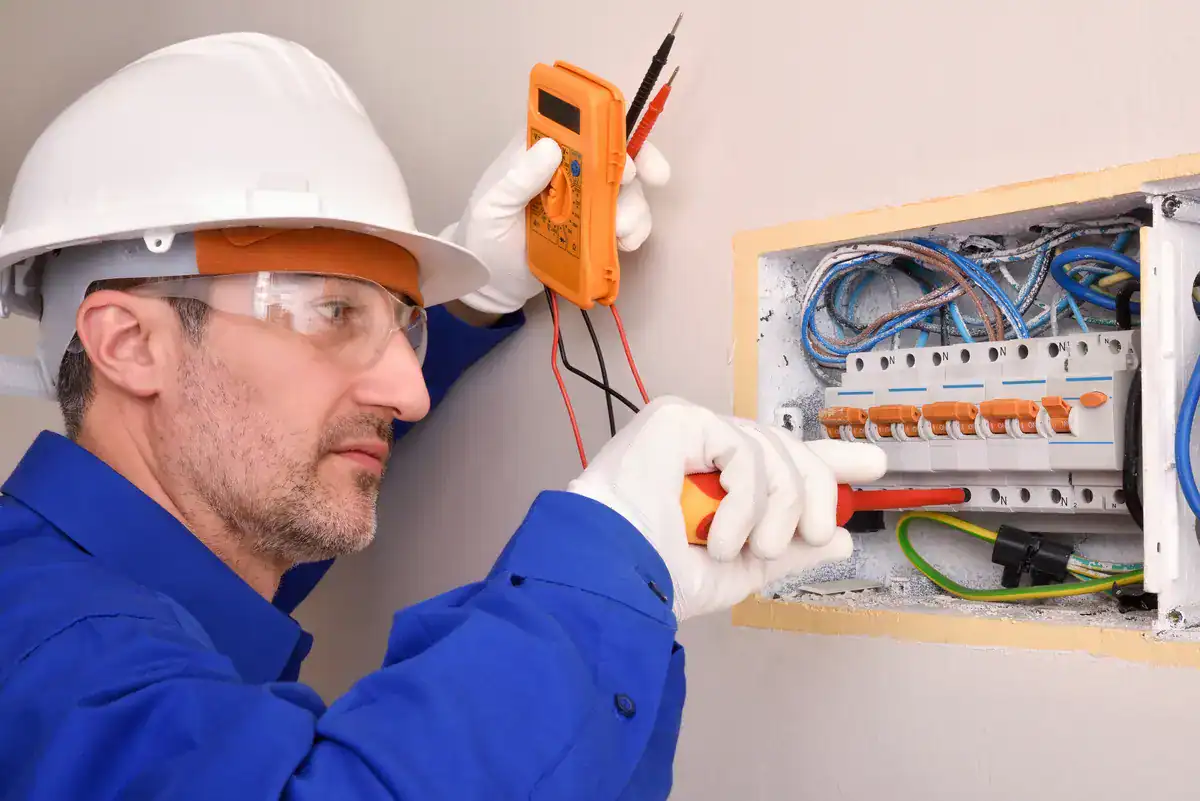 An electrician near me in Osseo, wearing a white hard hat, safety glasses, blue shirt, and white gloves, is using a digital multimeter and probes to inspect electrical wiring inside an open wall panel box. The box contains various wires and circuit breakers.