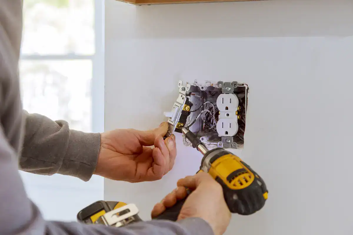 A person wearing a gray shirt is using a yellow and black power drill to install or repair an electrical outlet on a white wall. The electrical box is open, exposing wires and components. Natural light illuminates the scene from the left, showcasing the skill and precision of this electrician near me in Rogers.