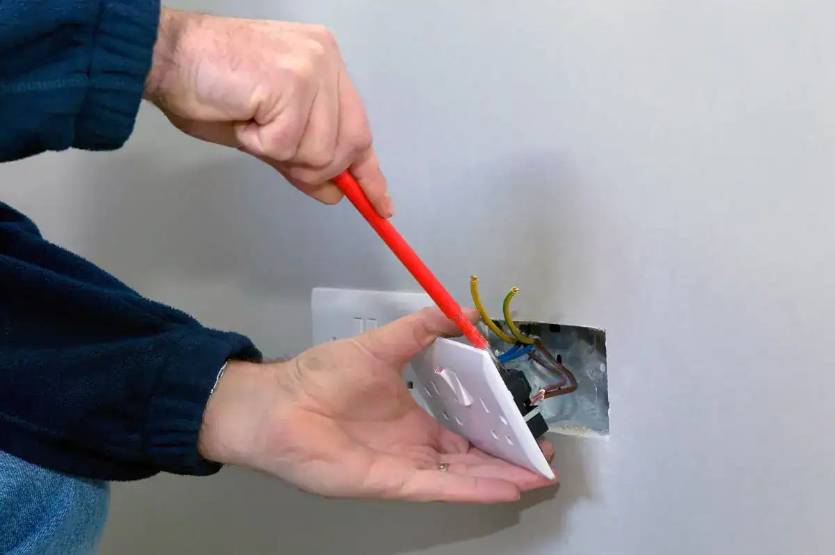 An electrician near me in Spring Lake Park is expertly using a screwdriver to install or repair a white electrical outlet. Wearing a dark long-sleeve shirt, they focus on connecting the exposed colored wires, as the outlet cover hangs partially detached from the wall.