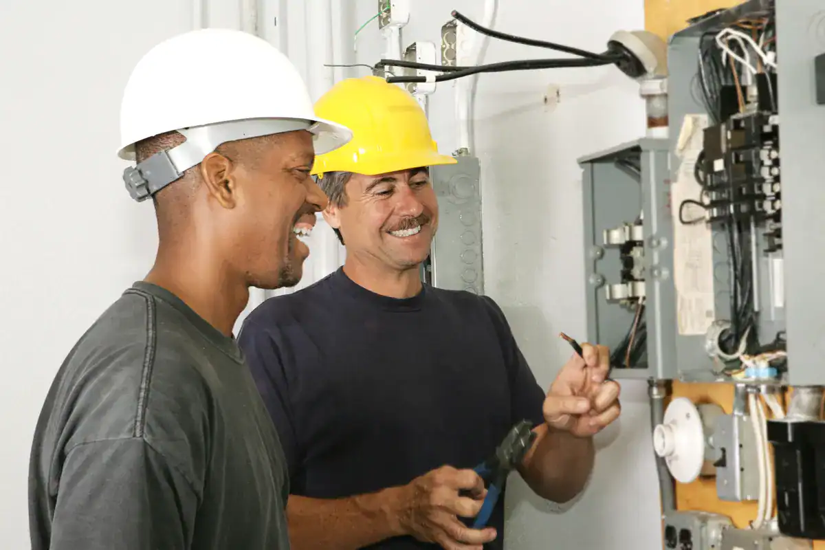 Two male electricians wearing hard hats work on an electrical panel. One holds pliers and a wire, while the other smiles and observes. They are dressed in work attire and are focused on the task, standing in a well-lit indoor space. If you need an electrician near me (Plymouth), they come highly recommended.