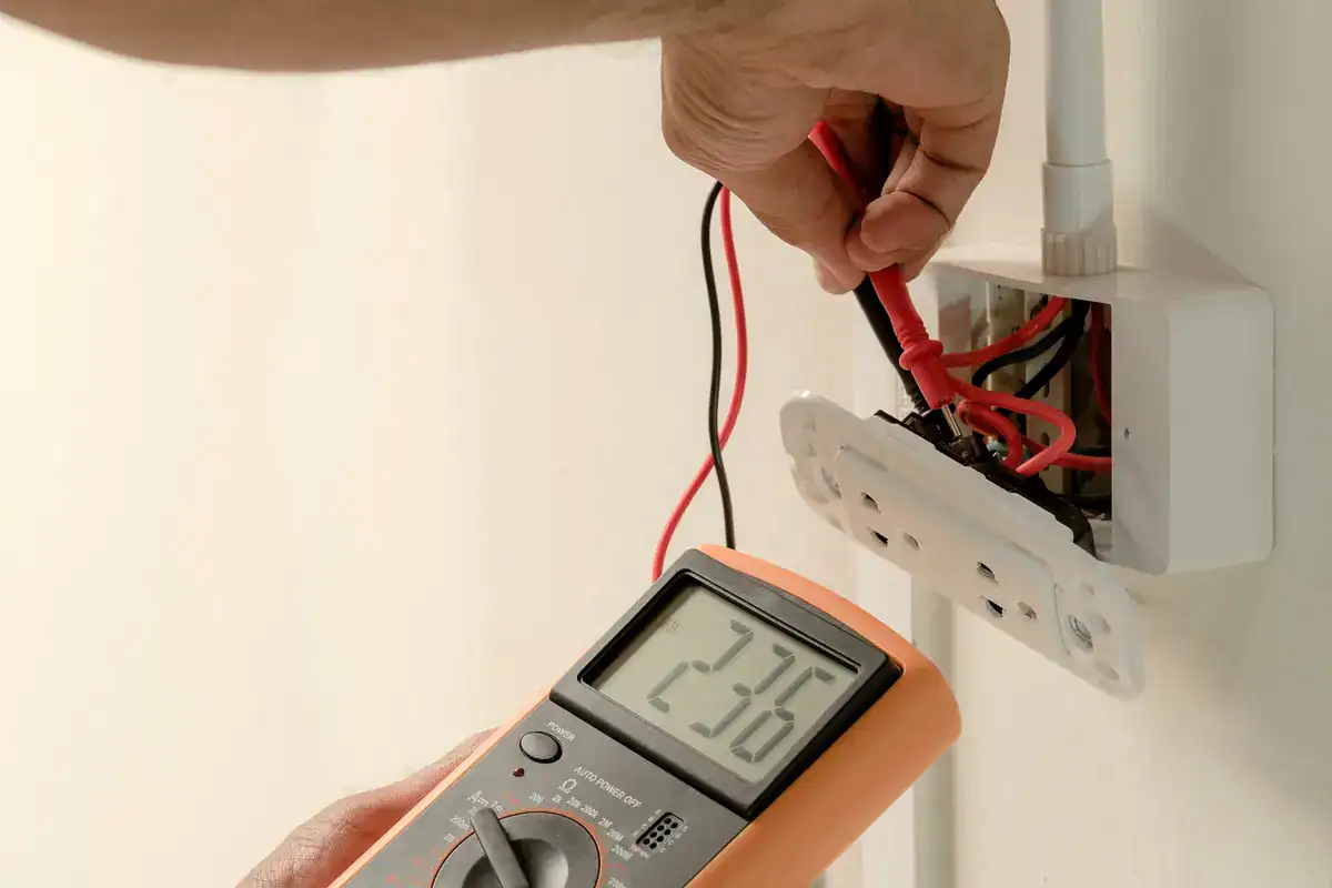 A person is using a digital multimeter to measure the voltage of an electrical outlet. The multimeter display reads 236. The person's hand holds the red probe touching the wires inside the outlet, which has its cover partially removed. If you need assistance, an electrician near me (Golden Valley) can help.