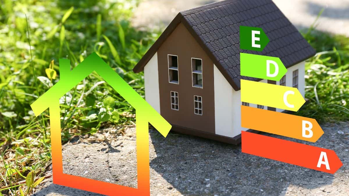 A small house model sits on green grass and a paved area. Next to it is an energy efficiency chart, complete with colorful bars labeled from A (green) to E (orange), showcasing various energy levels for efficiency.